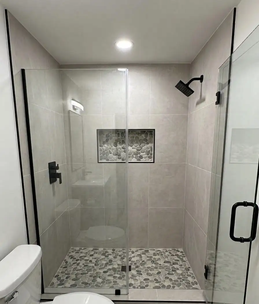 Bathroom remodeling Kent WA: walk-in shower behind a frameless glass door, gray porcelain wall tile, pebble shower floor, recessed niche and matte black fixtures