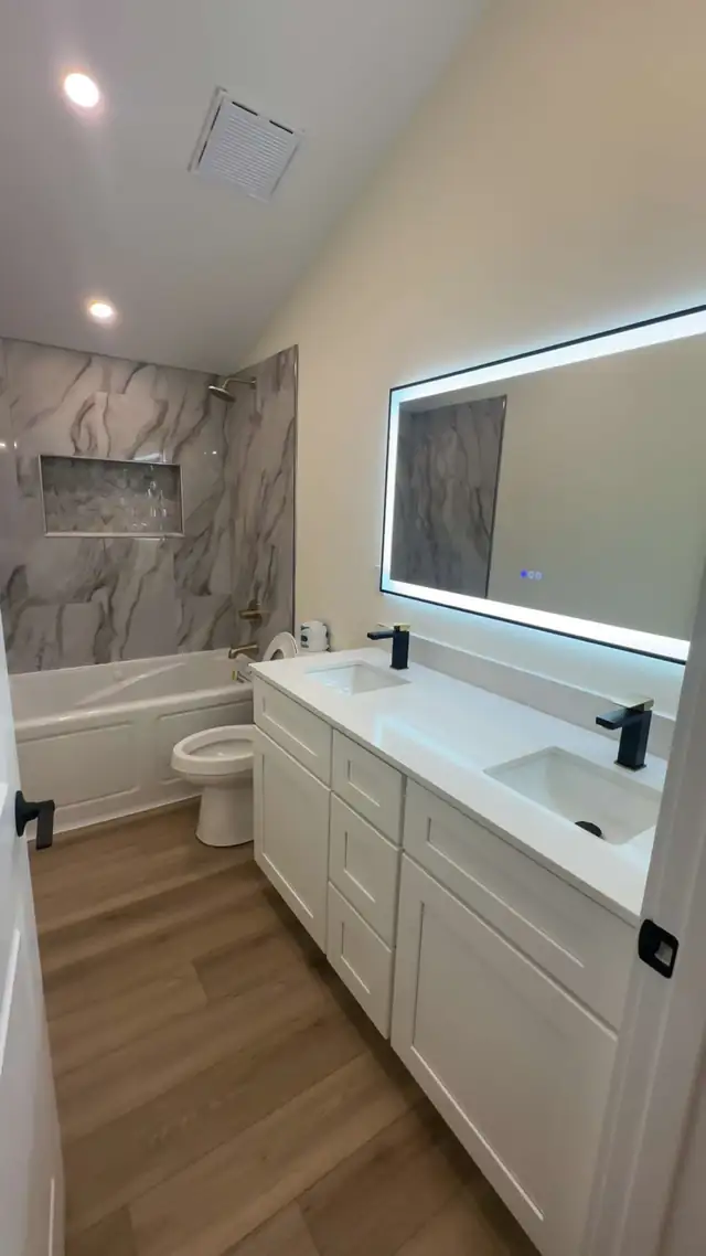Bathroom with white double vanity, backlit LED mirror, marble-look tub surround and wood-look LVP floor